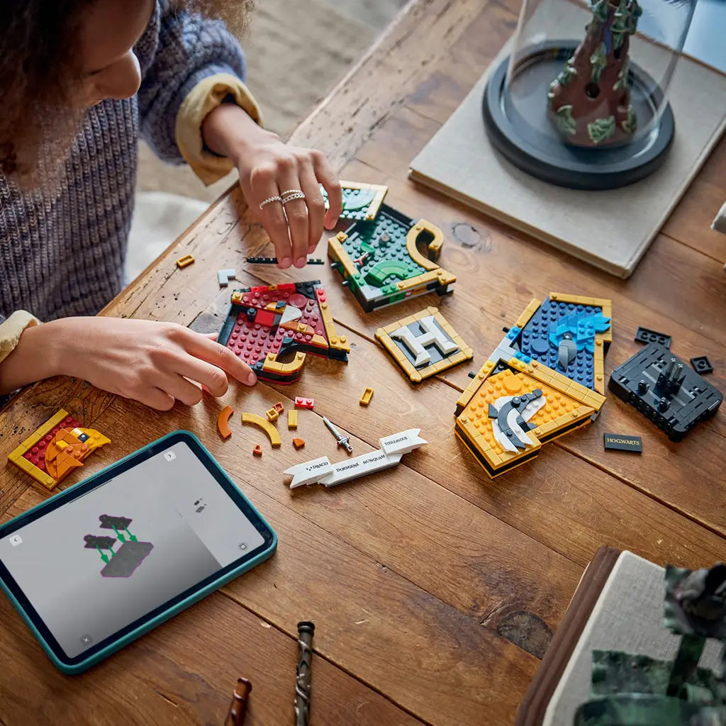 LEGO Harry Potter Hogwarts House Crest assembly in colorful plastic bricks, spread across a wooden table as a builder joins sections beside a tablet showing digital instructions.