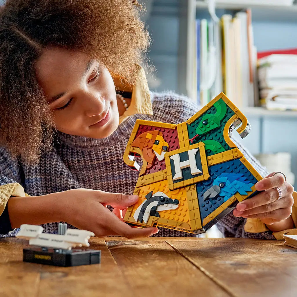 LEGO Harry Potter Hogwarts House Crest in colorful plastic bricks, held by a builder at a wooden table, with lion, snake, badger and eagle panels surrounding a central H.