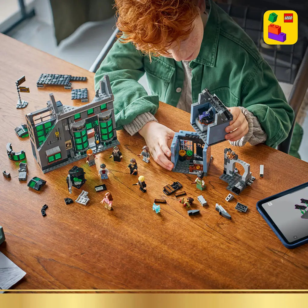 LEGO Harry Potter Knockturn Alley set in gray, black and green plastic bricks, with modular shops and minifigures being assembled on a wooden table beside a tablet.