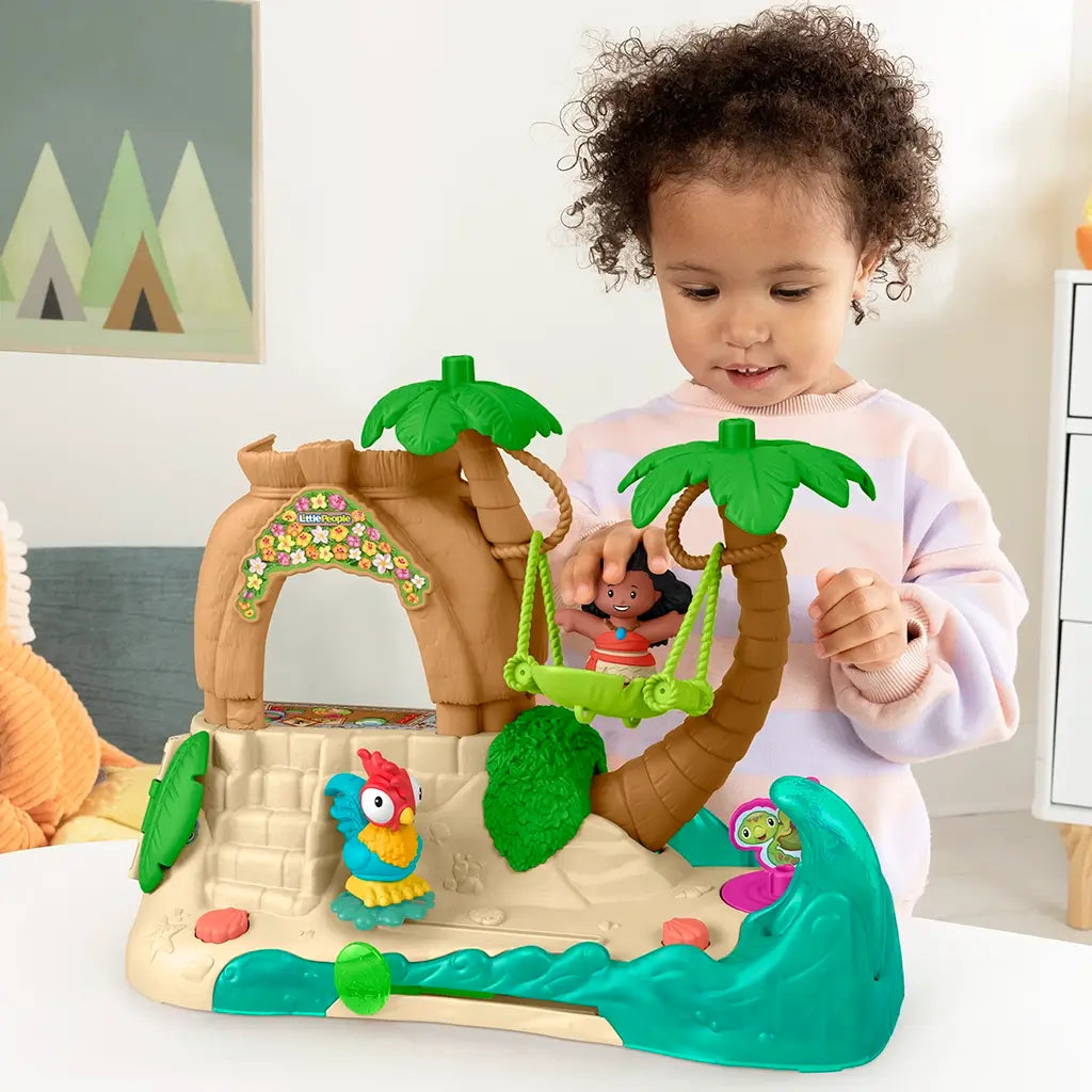 Smiling toddler interacting with Fisher-Price Little People Disney Moana Adventure Village playset, swinging Moana figure between palm trees on a tropical island-themed base.