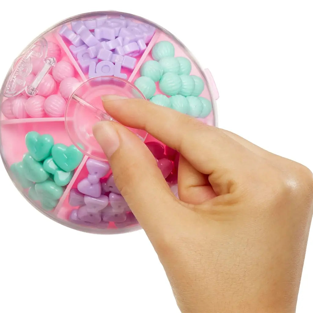 Overhead view of pink LOL Surprise Hair Beads storage ball divided into sections of pastel beads in purple, mint, pink, and heart shapes, with hand lifting the lid.