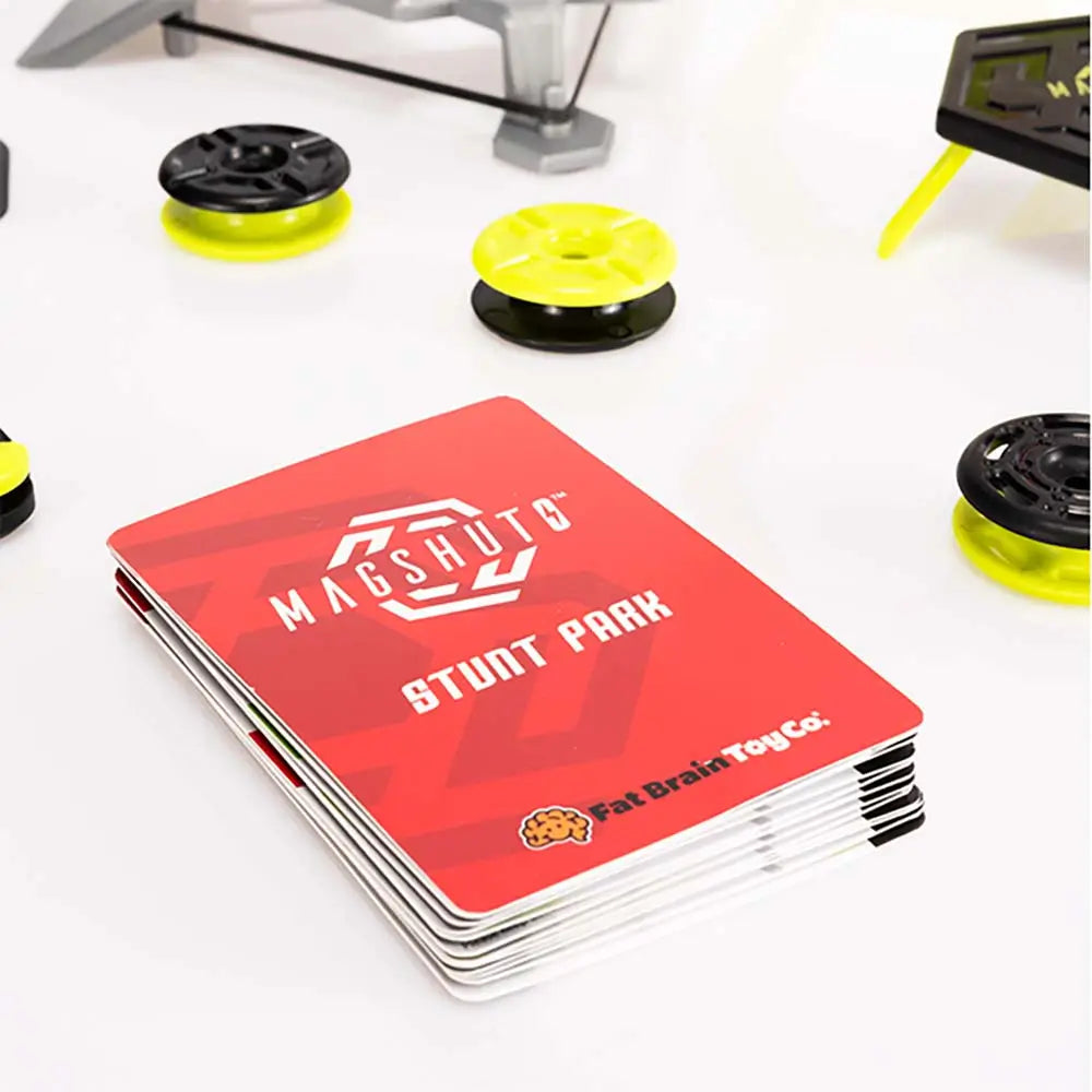 Magshuto Stunt Park challenge booklet with bold red cover and white logo lying on a smooth white surface, surrounded by scattered black and neon yellow magnetic discs and gray stunt park pieces.