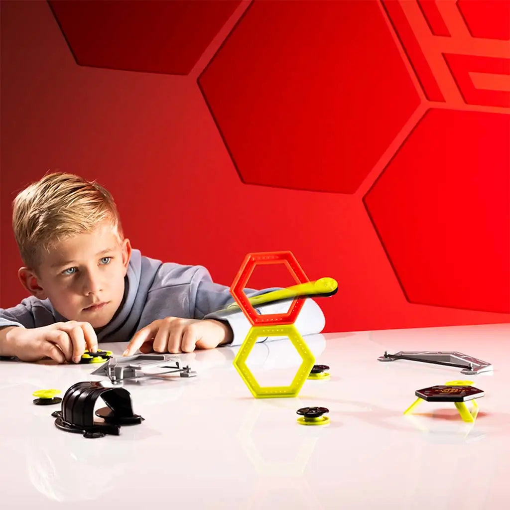 Focused child playing with the Magshuto Stunt Park set at a glossy white table, aiming neon yellow discs toward stacked red and yellow hexagon targets against a dramatic red geometric studio backdrop.