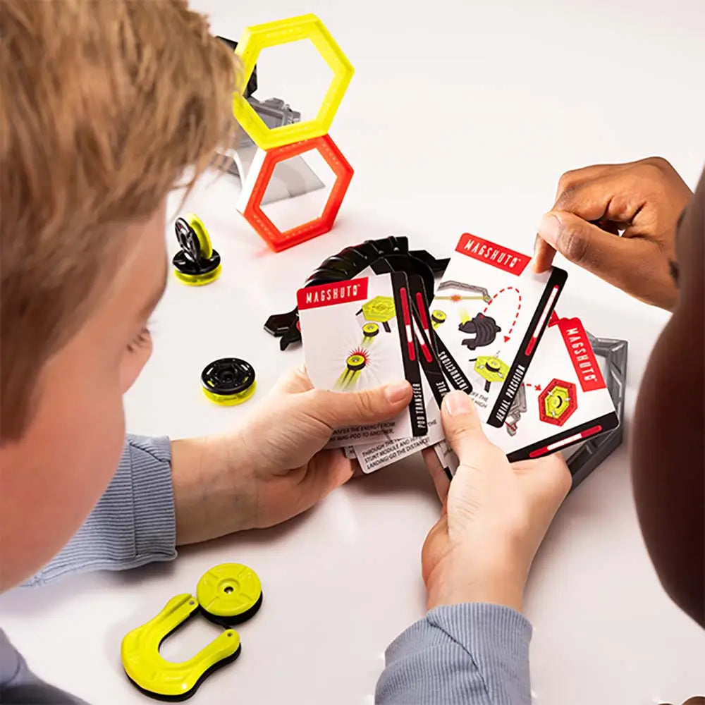 Two kids’ hands holding Magshuto Stunt Park challenge cards over a white table, with neon yellow discs, ramps, and hexagon targets scattered between them as they plan the next magnetic trick shot.