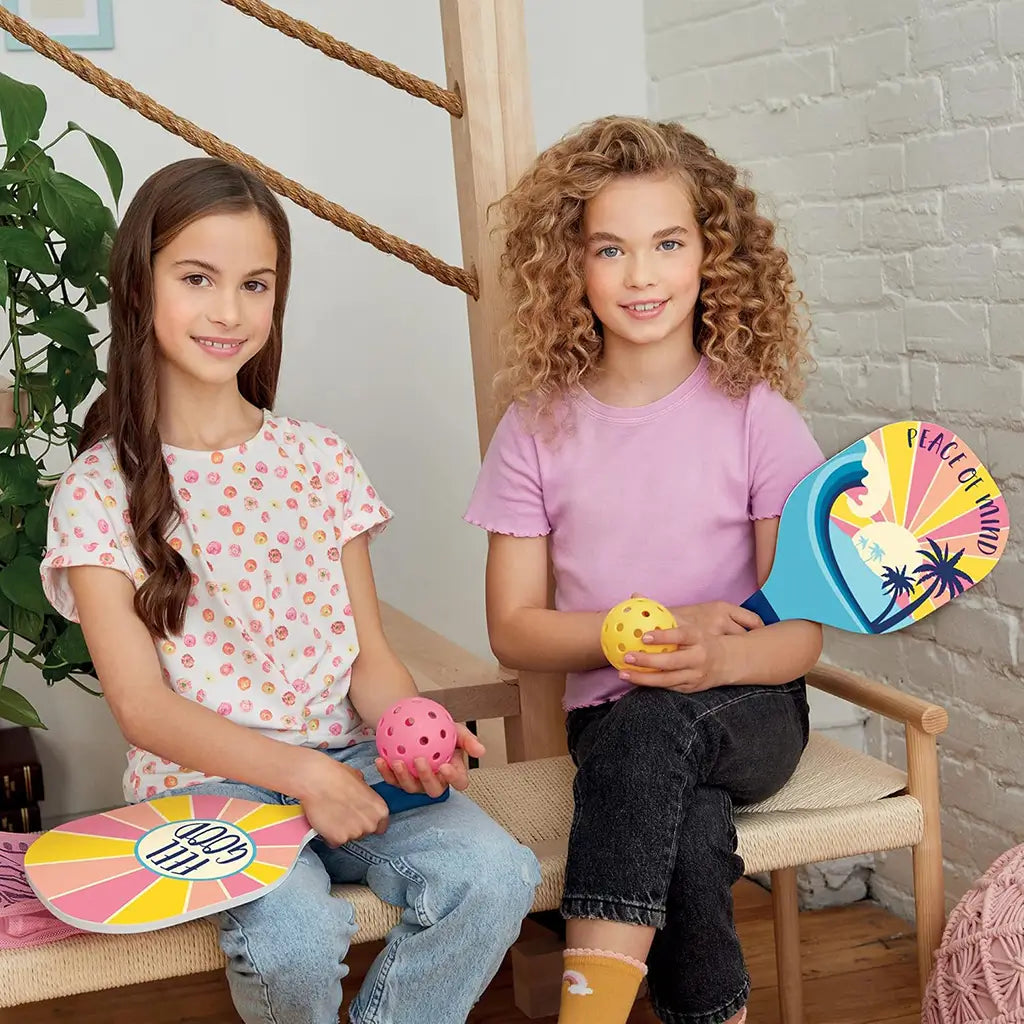 Two smiling kids sitting indoors holding colorful pickleball paddles and balls from the 3C4G Adventure Fun set, featuring bright “Feel Good” and “Peace of Mind” paddle designs.