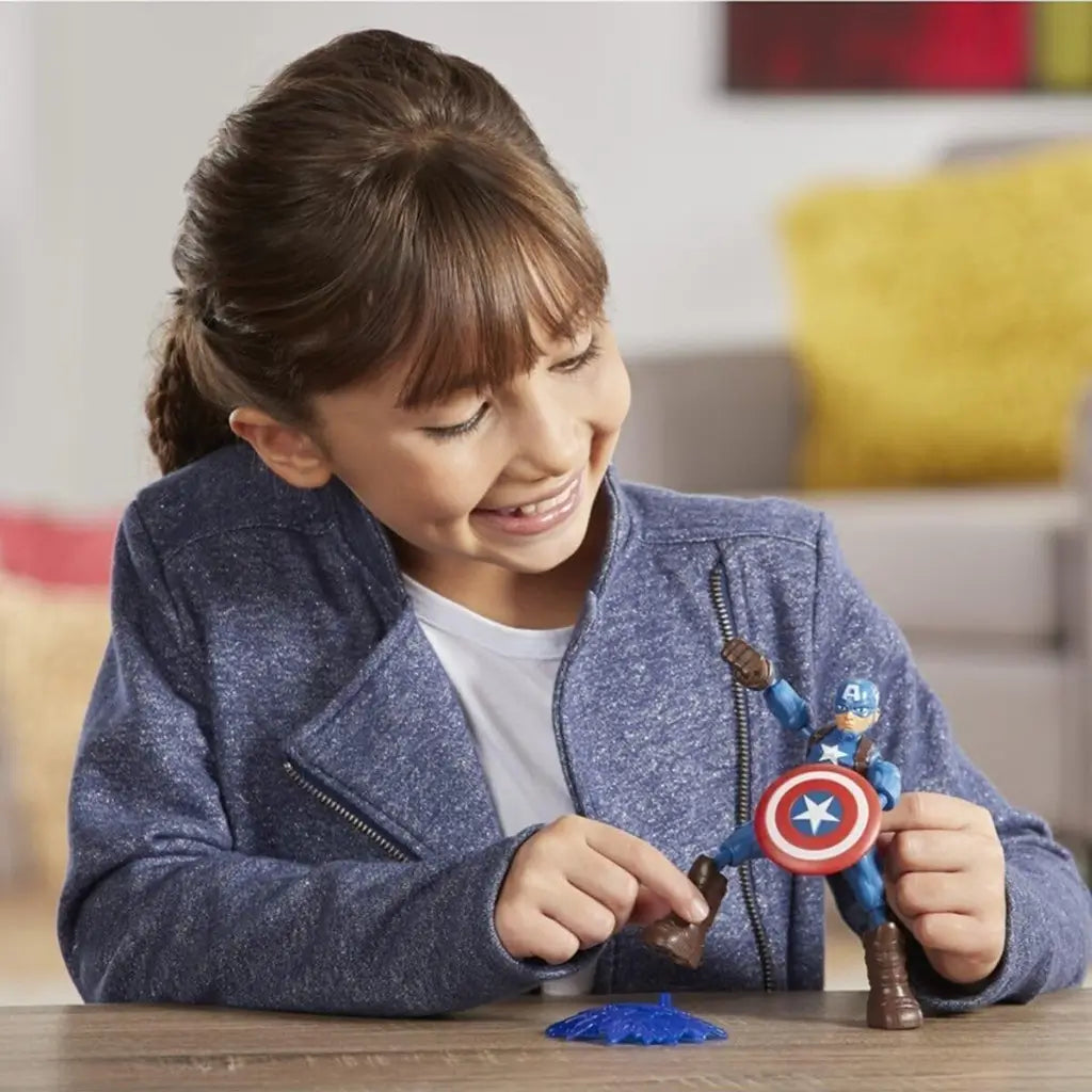 Smiling child in a blue jacket playing indoors with a Marvel Captain America Bend and Flex action figure, adjusting the toy’s pose and shield on a wooden table in a bright living room.