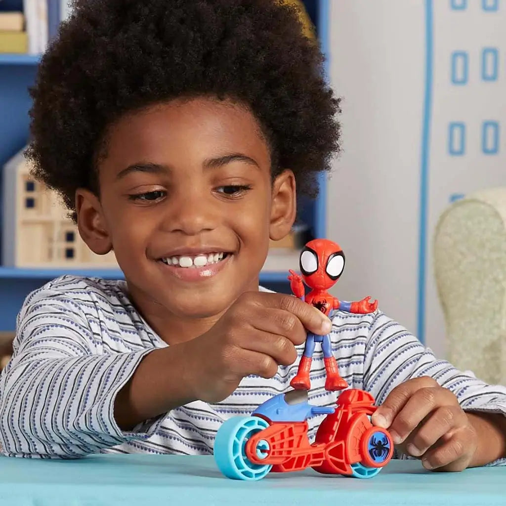 A smiling child plays with the Marvel Spidey motorcycle toy, holding the red-and-blue figure atop the matching bike with light blue wheels on a tabletop in a colorful indoor playroom setting.