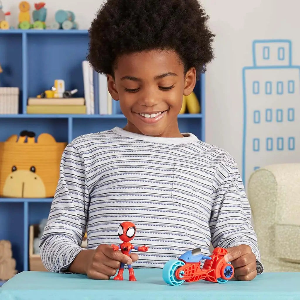 A cheerful child displays the Marvel Spidey figure and red-and-blue motorcycle with light blue wheels on a table in a bright playroom decorated with shelves and toys encouraging imaginative play.