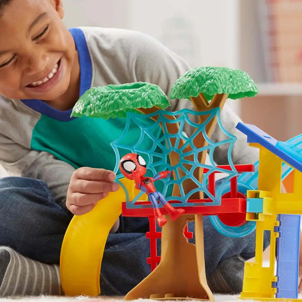 Marvel Spidey playground playset lifestyle scene shows smiling child guiding small Spidey figure across blue web lattice between tree canopy and yellow house, emphasizing large, chunky preschool design.