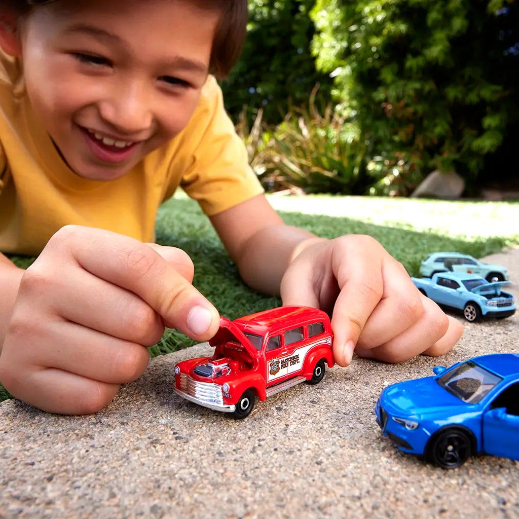 Smiling boy in yellow shirt plays outdoors with Matchbox diecast cars, holding a red fire rescue truck with opening hood while blue and silver cars sit nearby on pavement near grass.