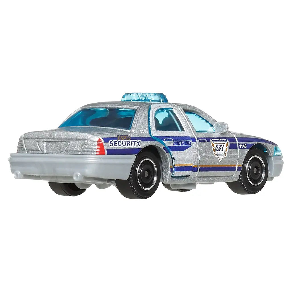 Rear angled view of Matchbox Ford Crown Victoria security car toy in silver with blue accents, showcasing tail lights, security decals, detailed trunk lines, and black wheels.