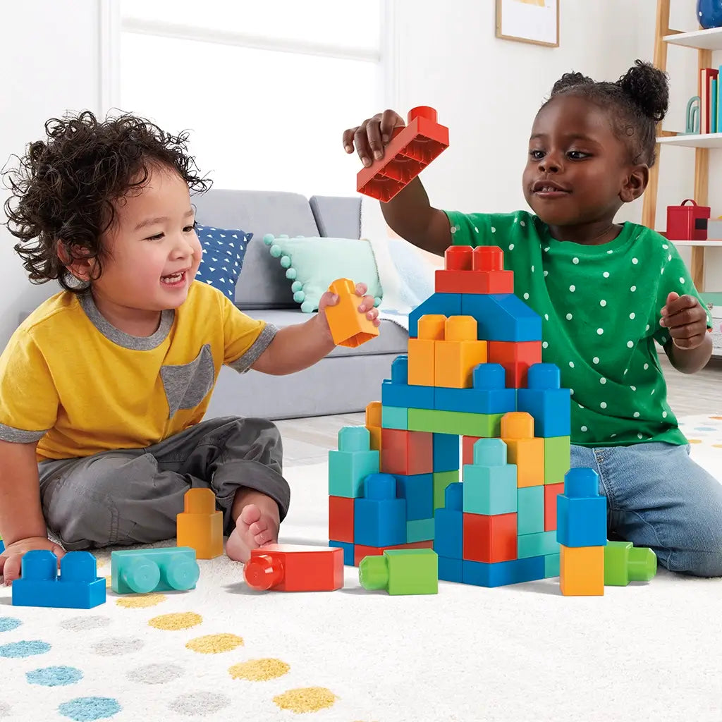 Two smiling toddlers sit on a carpeted floor building a colorful Mega Bloks tower with red, blue, green, and yellow plastic blocks in a cozy living room setting.