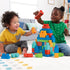 Two smiling toddlers sit on a carpeted floor building a colorful Mega Bloks tower with red, blue, green, and yellow plastic blocks in a cozy living room setting.