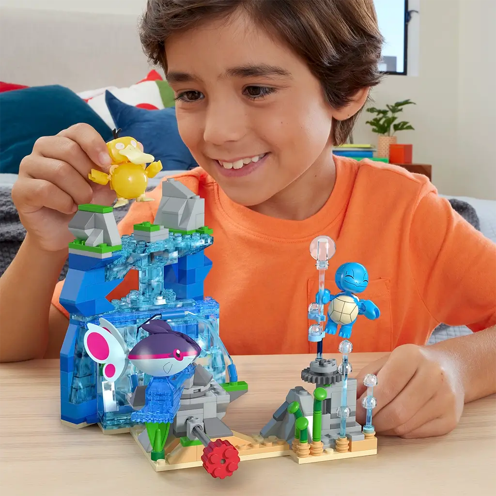 Child in orange shirt plays with Mega Pokémon Aquatic Adventure building set, featuring colorful water-themed blocks with Squirtle, Psyduck, and Finneon figures on a wooden table.