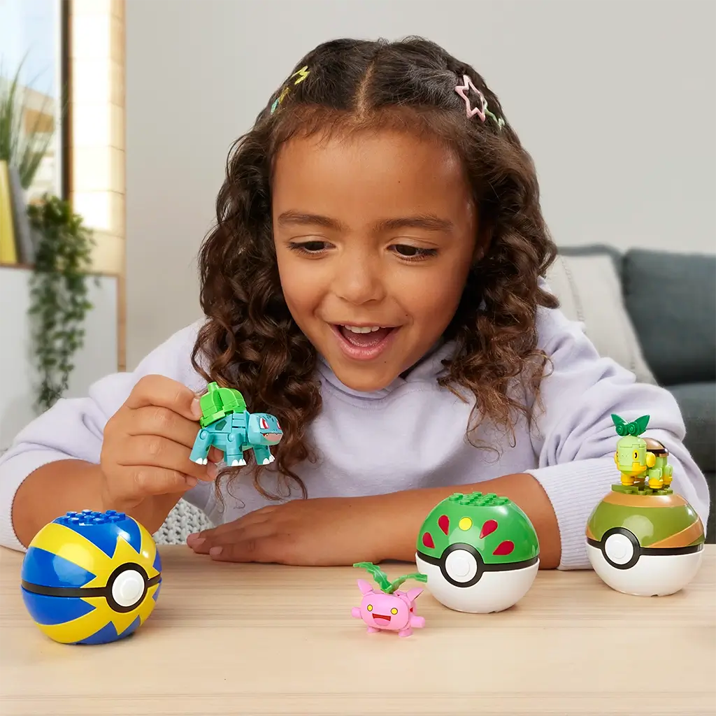 Smiling young girl plays with Mega Pokémon Bulbasaur figure alongside Hoppip, Turtwig, and colorful Poké Ball toys, showing creative building and interactive fun.