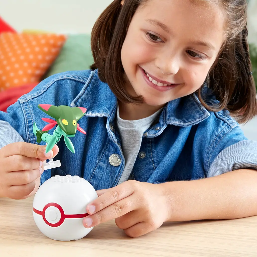 Smiling child in denim jacket holding Mega Pokémon Dreepy figure above white Poké Ball toy on wooden table in bright indoor playroom setting. This Mega Construx building set includes 21 pieces for creative play, recommended for Pokémon fans ages six and older.