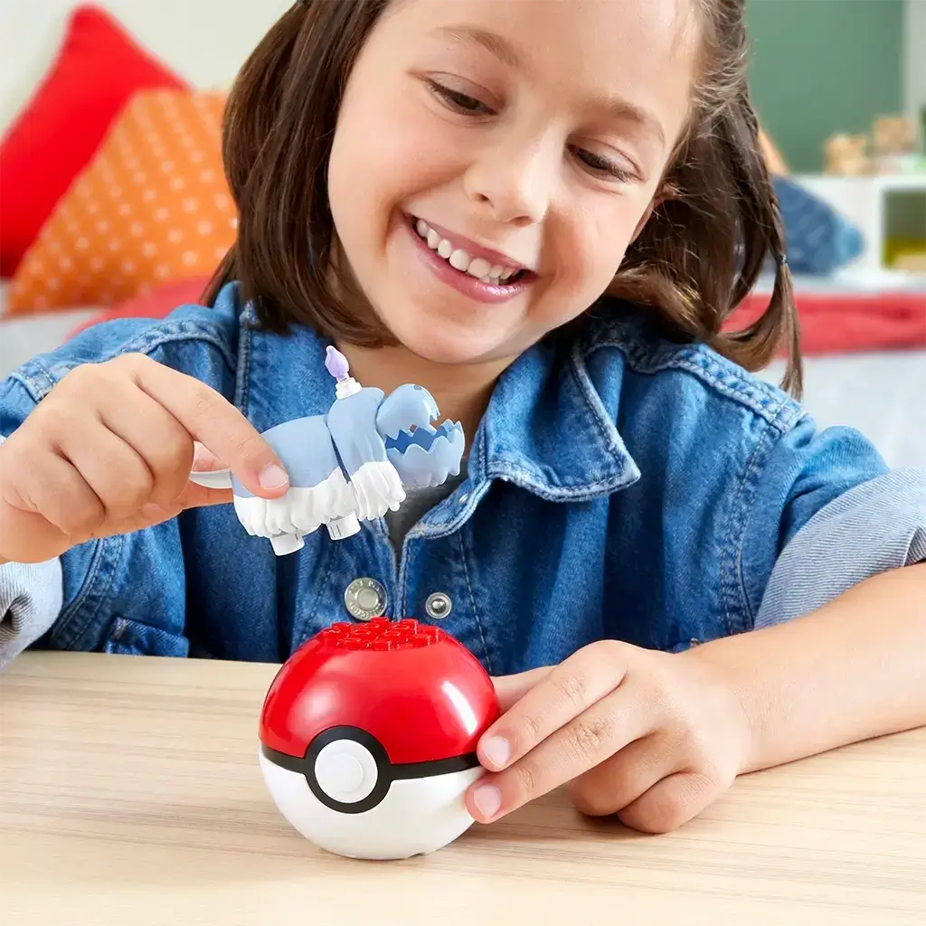 Smiling child in denim shirt placing Mega Pokémon Greavard figure into red and white Poké Ball toy while seated at wooden table in a bright room. This Mega Construx toy set offers build-and-play fun for Pokémon fans ages six and older, combining display and play features in one collectible set.