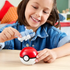 Smiling child in denim shirt placing Mega Pokémon Greavard figure into red and white Poké Ball toy while seated at wooden table in a bright room. This Mega Construx toy set offers build-and-play fun for Pokémon fans ages six and older, combining display and play features in one collectible set.