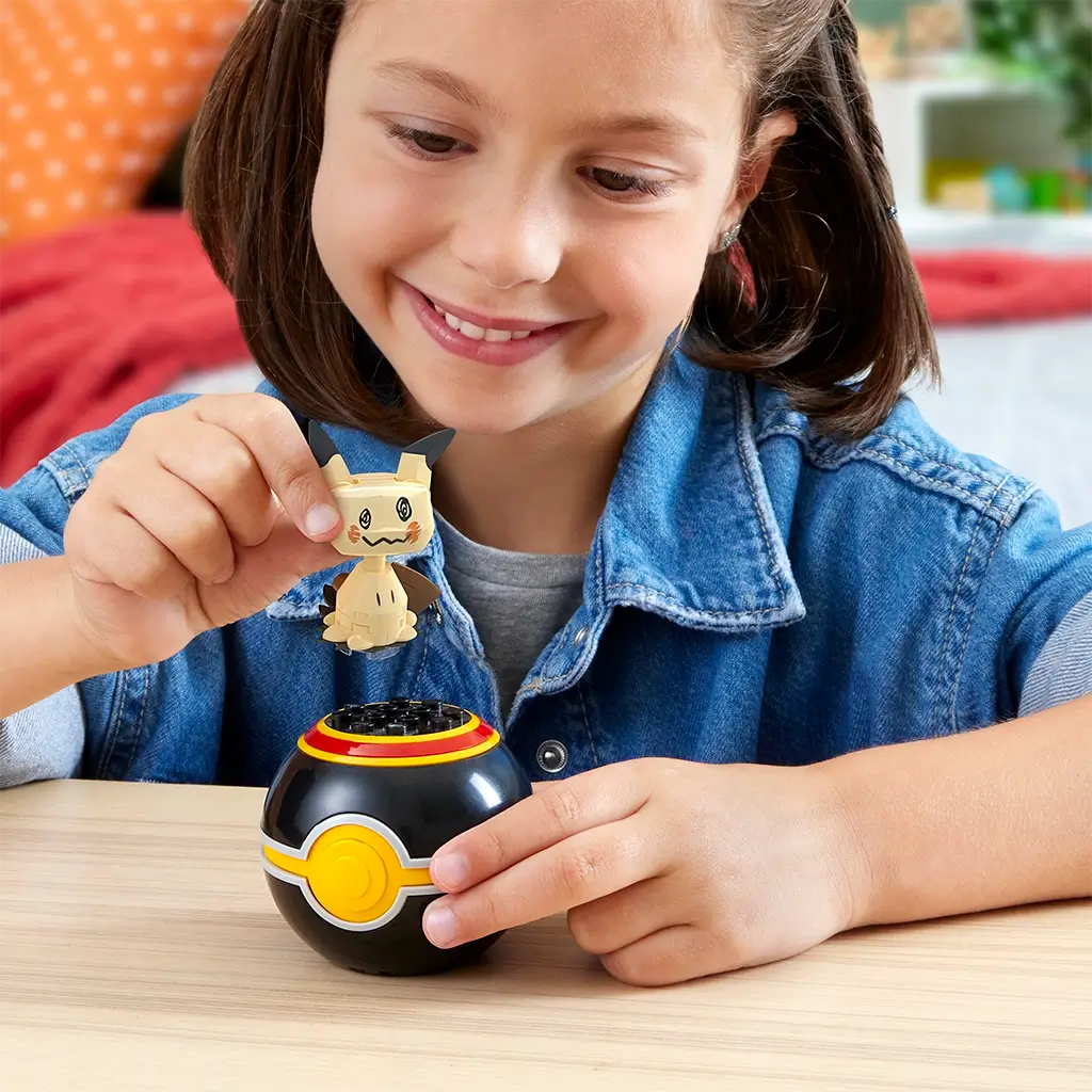 Smiling child in denim shirt playing with Mega Pokémon Mimikyu figure, placing it into the black Poké Ball toy on a wooden table in a bright indoor setting.