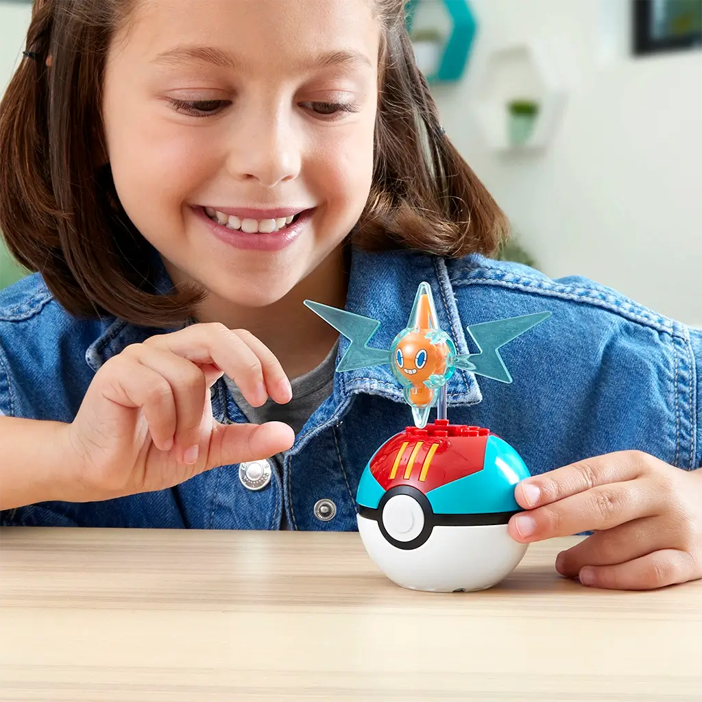 Smiling child in denim jacket playing with MEGA Pokémon Rotom building set, featuring orange Rotom figure with translucent blue wings emerging from a red and blue Poké Ball.