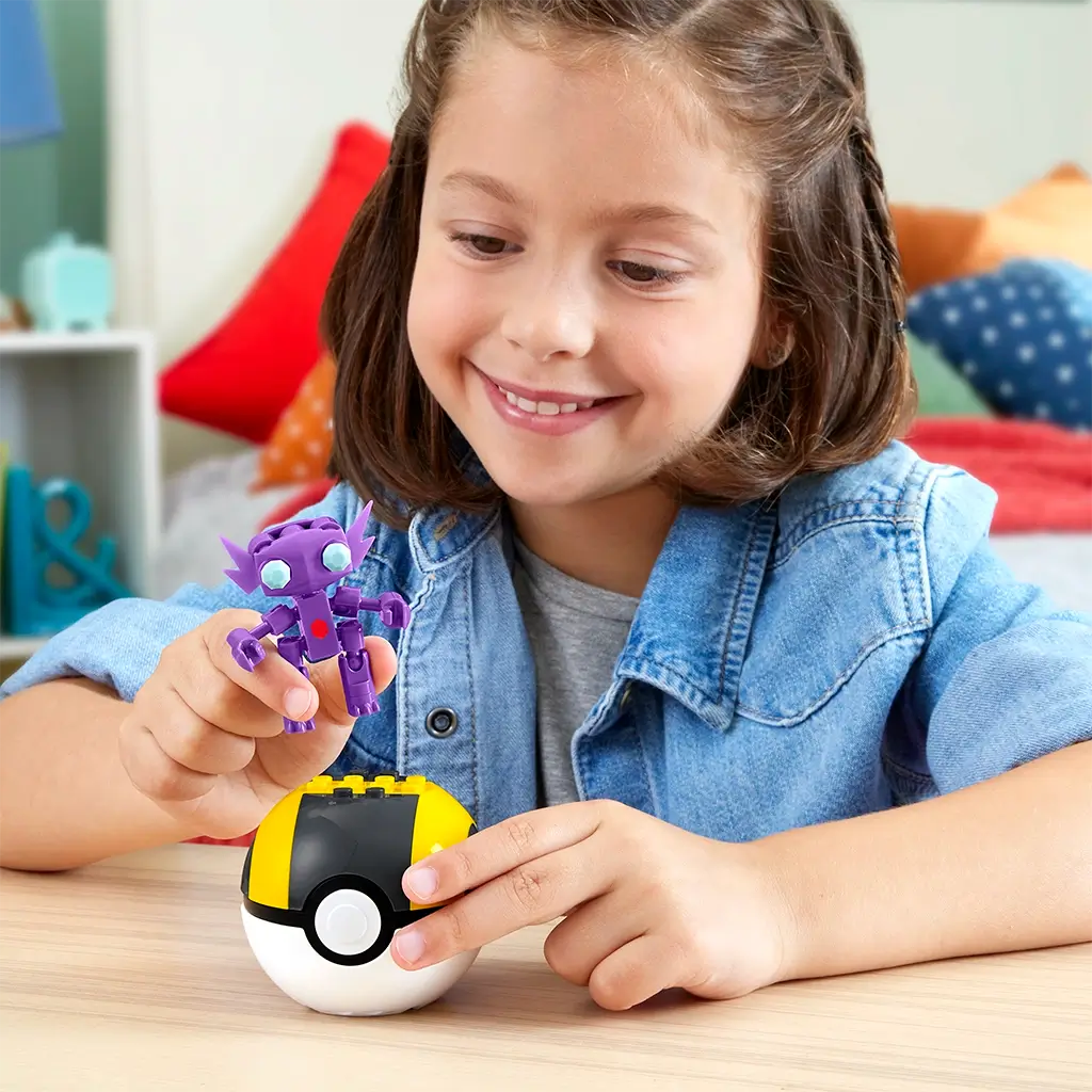 Child smiling while holding Mega Pokémon Sableye figure above yellow-and-black Ultra Ball building block case, showing imaginative play in a colorful bedroom setting.