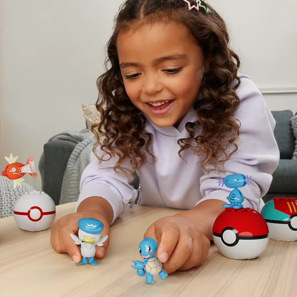 Young girl smiling and playing with MEGA Pokémon Water-Type Trainer Team figures, including Squirtle, Quaxly, Wooper, and Magikarp, each displayed with their respective Poké Balls.