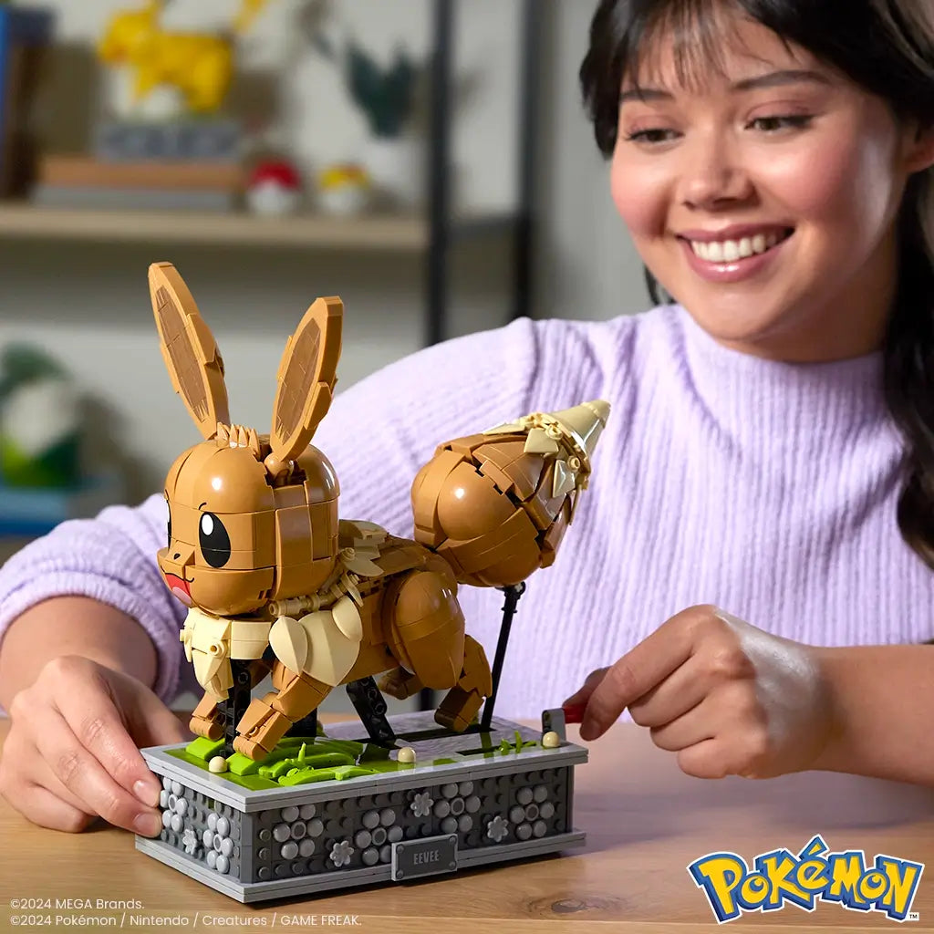 Smiling woman in lavender sweater turning crank on brown Mega Pokémon Eevee building set, displayed on a gray floral-patterned base with green grass accents.