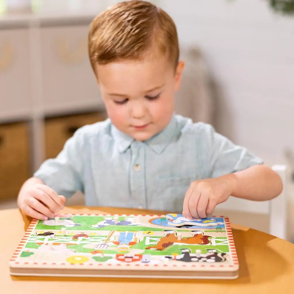 A young boy in a light blue shirt sits at a wooden table, concentrating as he places pieces into the Melissa & Doug Old MacDonald’s Farm sound puzzle, enhancing fine motor and problem-solving skills.