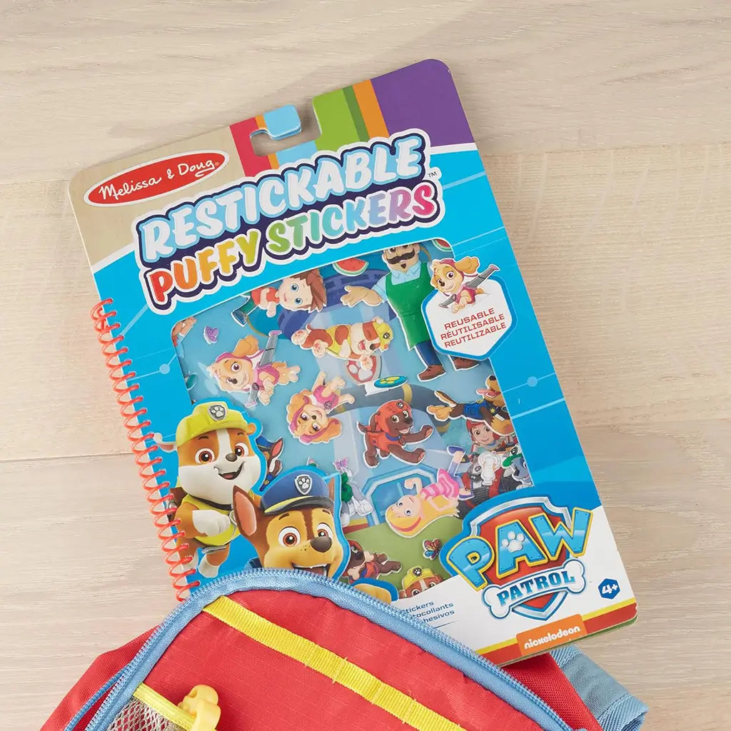 Paw Patrol puffy sticker set partially tucked into a red and yellow backpack, showing Melissa & Doug packaging with bright characters and reusable stickers for imaginative play.