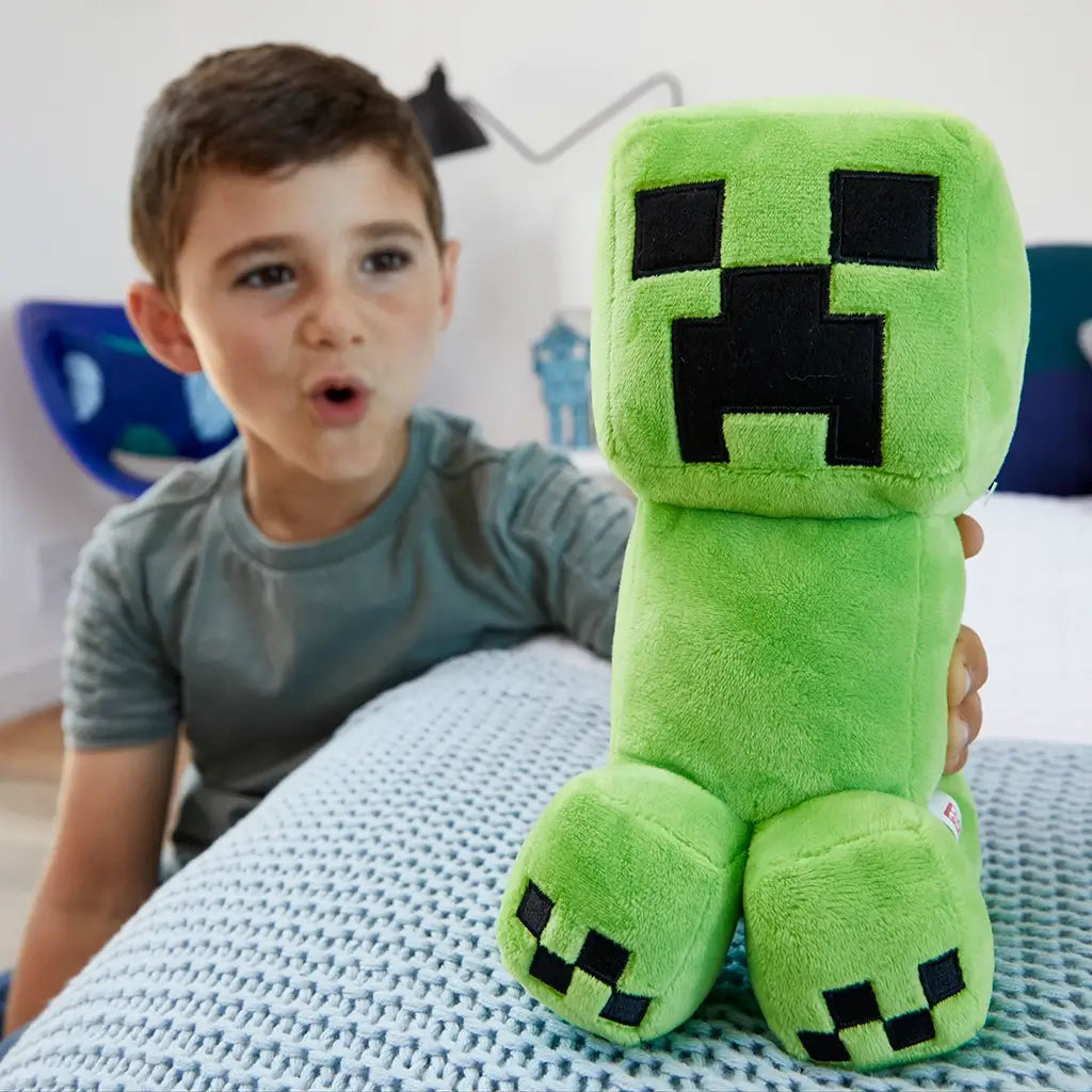 Minecraft Creeper plush in bright green soft fabric with pixelated black face, blocky shape and short legs, shown in a lifestyle bedroom setting with a child holding the toy on a bed.