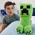 Minecraft Creeper plush in bright green soft fabric with pixelated black face, blocky shape and short legs, shown in a lifestyle bedroom setting with a child holding the toy on a bed.