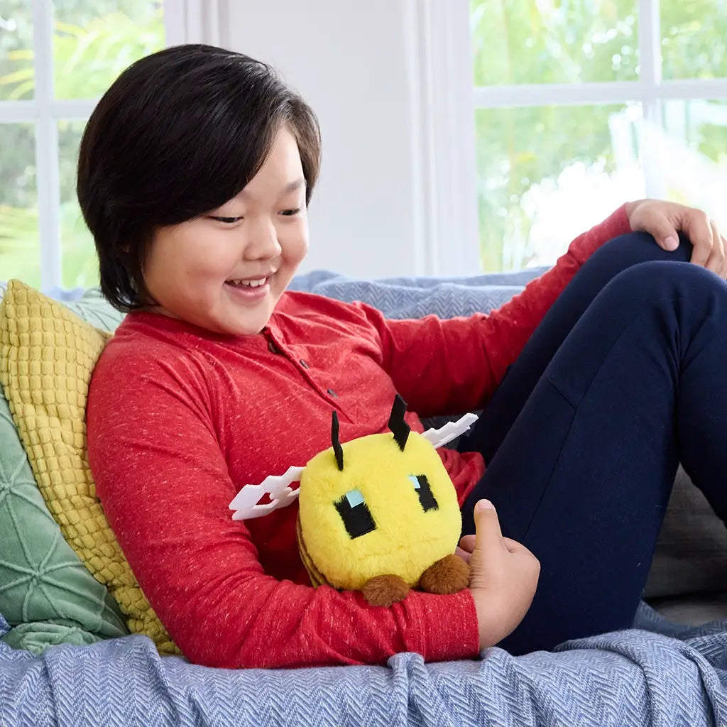 Smiling child in red shirt sits on a sofa holding the soft Minecraft bee plush, showing yellow blocky design with wings and antennae in cozy living room setting.