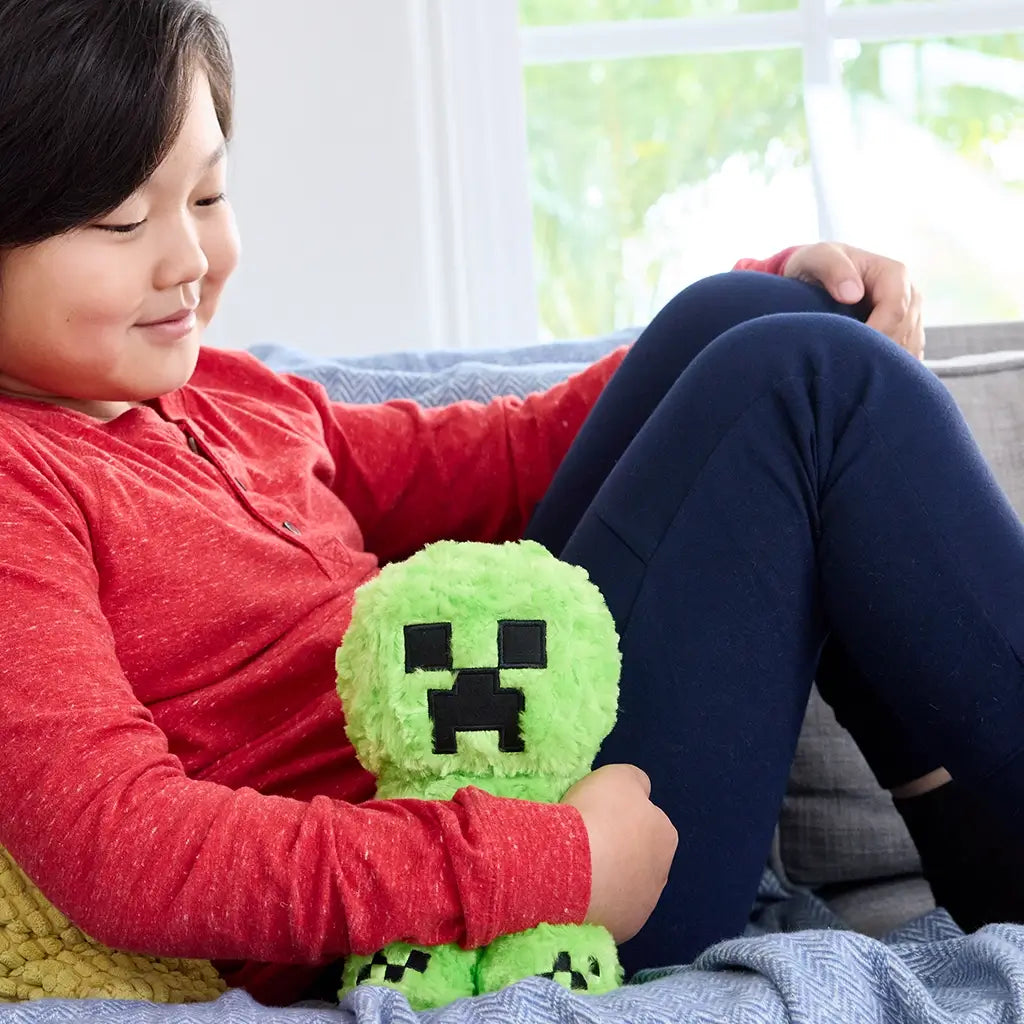 Child wearing a red shirt smiles while holding a fuzzy green Minecraft Creeper plush toy with pixelated black stitched face, sitting indoors on a sofa.