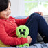 Child wearing a red shirt smiles while holding a fuzzy green Minecraft Creeper plush toy with pixelated black stitched face, sitting indoors on a sofa.