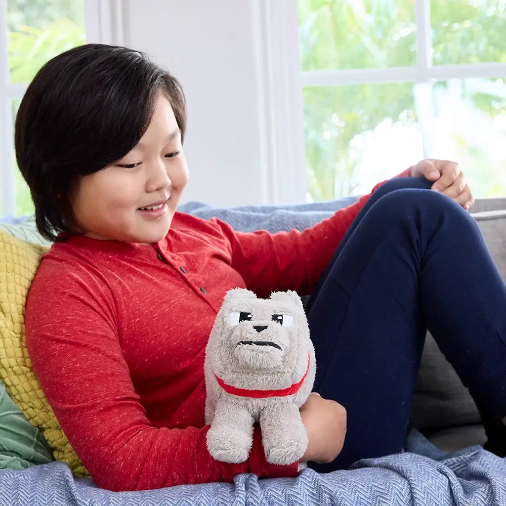 Child in red shirt smiles on couch while holding Minecraft Dennis plush wolf, highlighting soft beige fur, red collar, blocky cube-shaped design, and screen-accurate embroidered face.