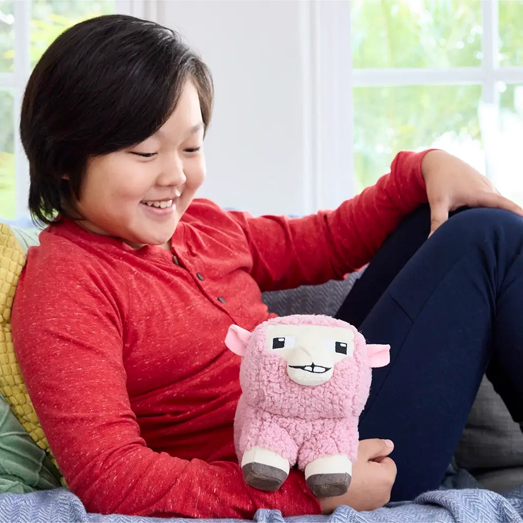 Child smiling while holding Minecraft Movie pink sheep plush, showing the 8-inch toy’s fluffy pink fleece, square face with embroidered eyes, and blocky legs, sitting indoors on a gray sofa.