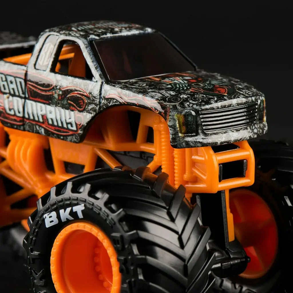 Close-up of Monster Jam Bad Company 1:64 monster truck, emphasizing detailed skull and flame graphics, orange suspension frame, and aggressive treaded wheels.