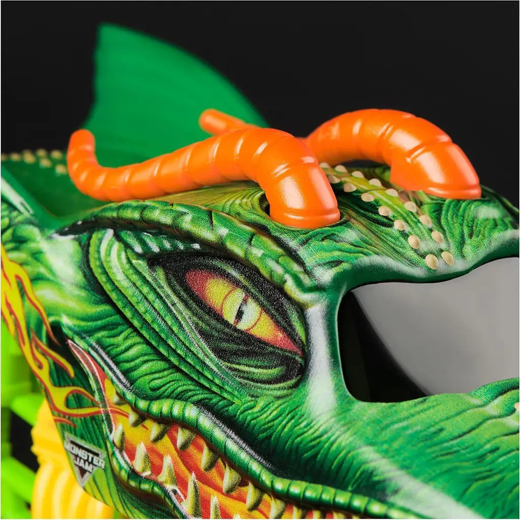 Closeup of Monster Jam Dragon diecast truck shows green dragon face with textured scales, fiery eyes, sharp teeth, and bright orange horns against a glossy black window background.