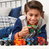 Monster Jam toy trucks in multicolor plastic with large rubber wheels held by a smiling child playing on a bedroom floor setting with bed and pillows in the background.