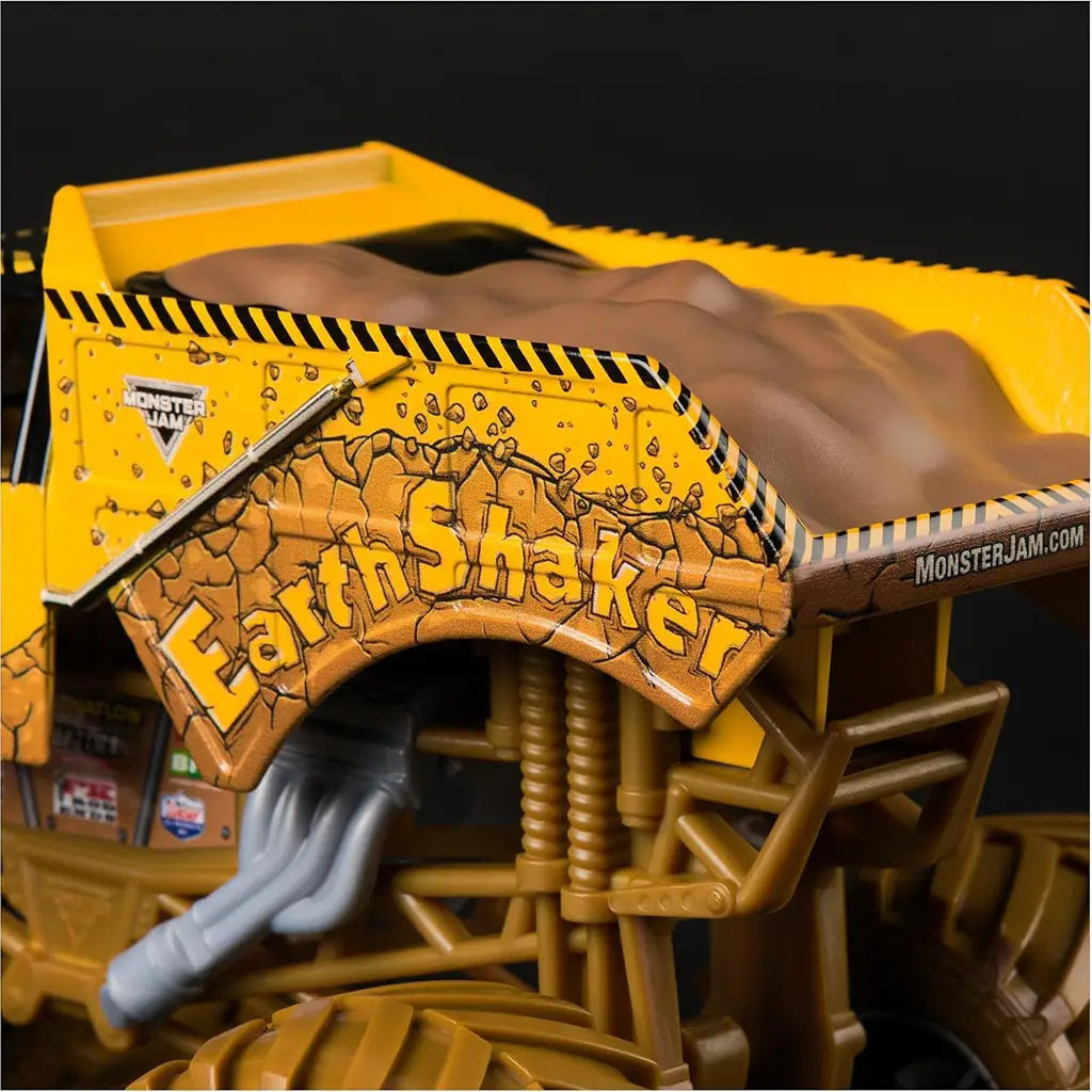 Monster Jam Earth Shaker diecast truck closeup shows rugged yellow body with cracked rock graphics, raised dirt bed, and detailed suspension pipes against a dark background.
