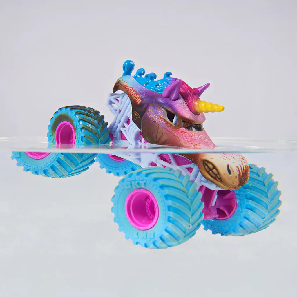 Monster Jam Sparkle Smash toy truck in pink, blue, and brown plastic with mud texture, teal tires and pink rims, partially submerged in water to demonstrate color change feature, set against a clean minimal background