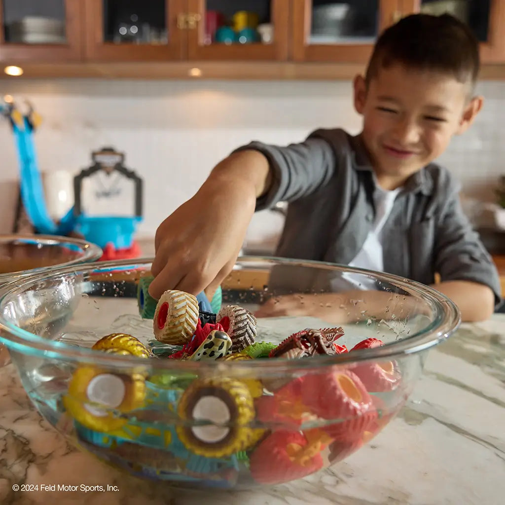 Monster Jam color change toy trucks in multicolor plastic with large textured wheels and rugged shapes, designed for water play, placed in a glass bowl with a smiling child interacting in a bright kitchen background