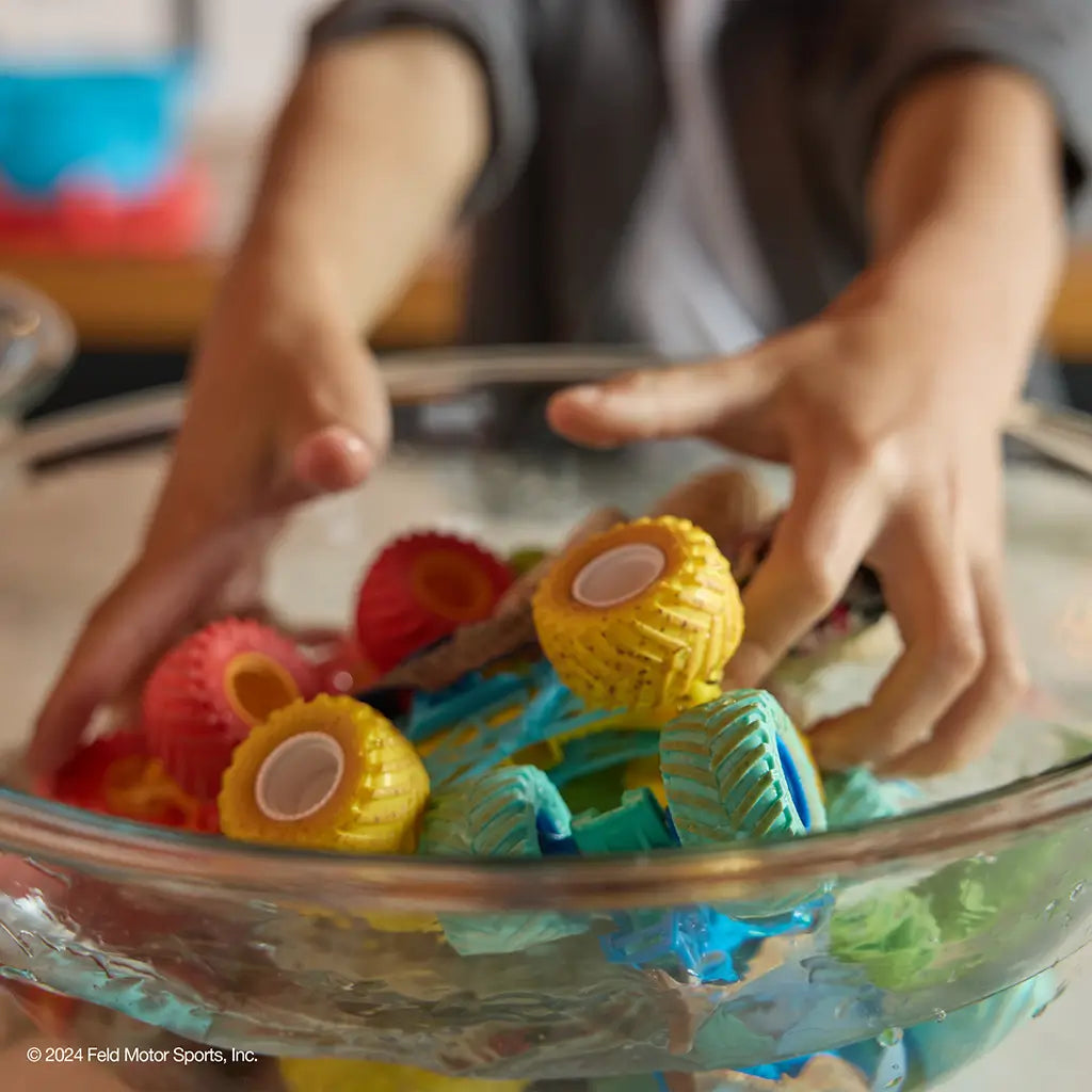 Monster Jam color change toy trucks in multicolor plastic with textured tires and chunky forms, designed for water play transformation, shown inside a clear bowl with hands interacting in a bright indoor setting