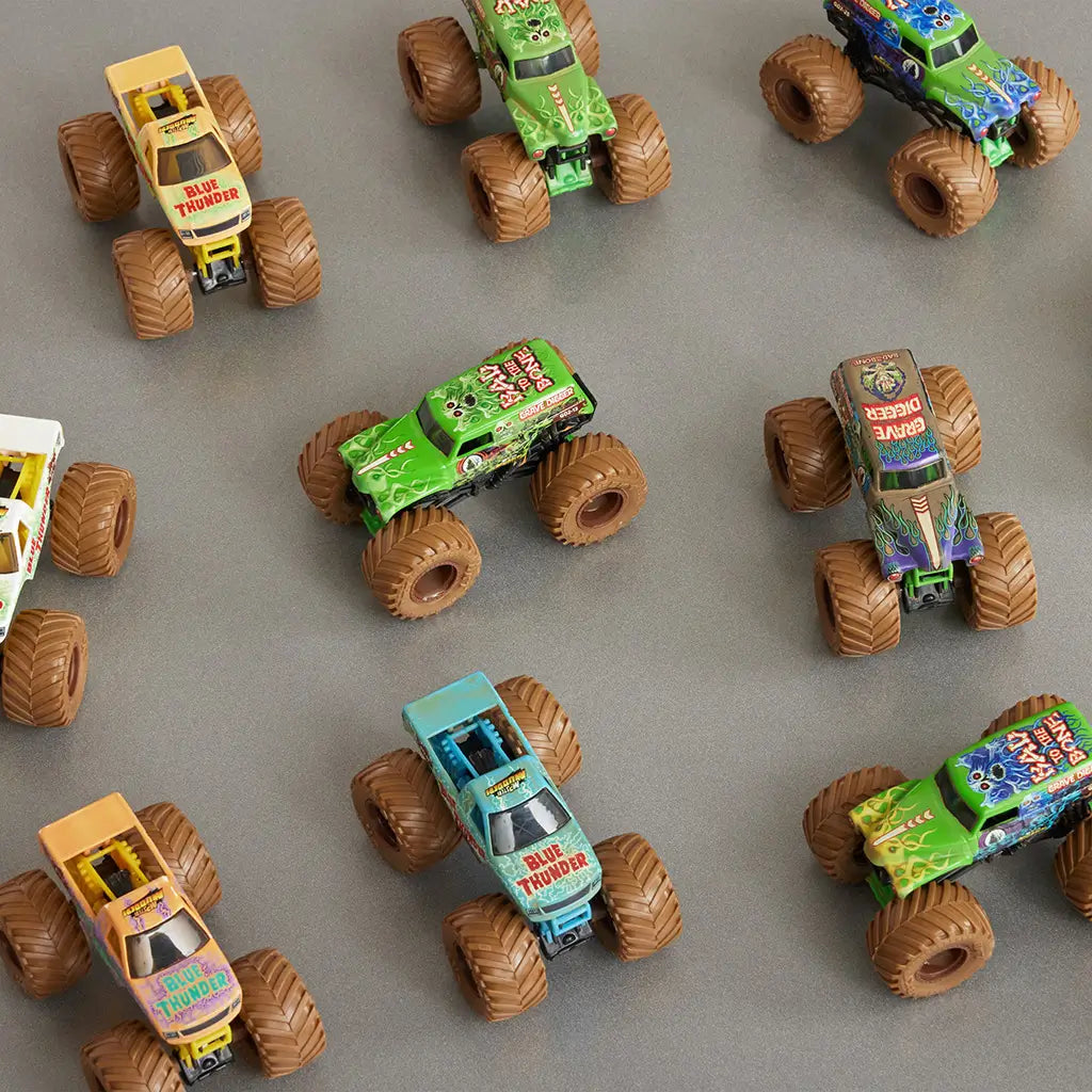 Monster Jam Mystery Mudders toy trucks in multicolor plastic with muddy textured tires, oversized monster truck shapes for collection, scattered across a flat tabletop surface in a display setting.