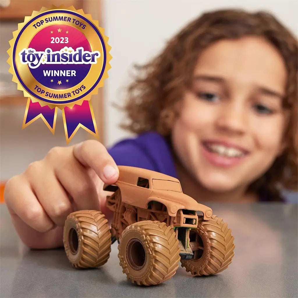 Monster Jam toy truck coated in brown mud with plastic body, oversized rubber tires, and compact shape, pushed by a child’s hand on a tabletop, featuring award badge graphic for top summer toy recognition.