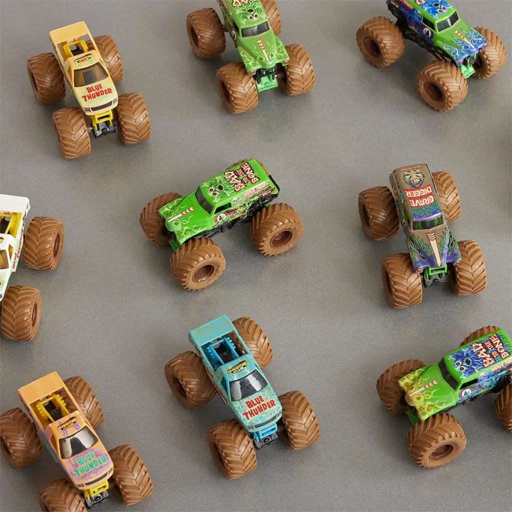 Monster Jam toy trucks in mixed bright colors with plastic bodies, thick rubber tires, and rectangular forms, arranged in a scattered top view layout on a smooth neutral tabletop for collectible display.