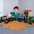 A smiling child plays with the Monster Jam Son-Uva Digger diecast truck on textured brown terrain, showcasing large BKT tires, blue chassis, and bold graphic bodywork in a cozy indoor playroom.