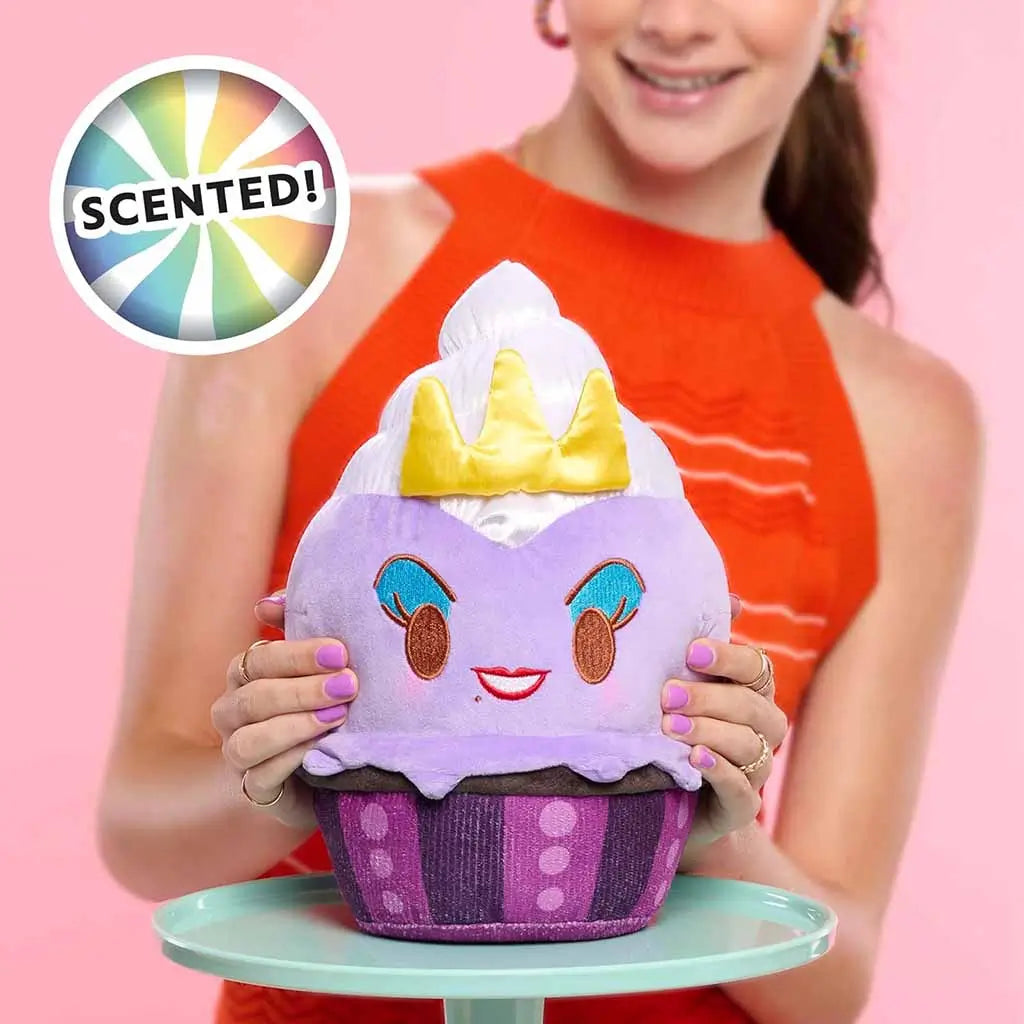 Close-up of a scented Disney Munchlings Ursula plush cupcake from The Little Mermaid, showing a purple base, white whipped top, yellow crown, and embroidered smiling face held by a person in red top.