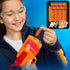 NERF N-Series dart blaster scene with a child in safety goggles loading a small orange foam dart into the bright orange and red toy, while an inset graphic highlights the included twelve refill darts.