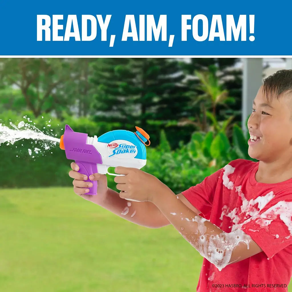 Smiling boy in red shirt spraying foam with a Nerf Super Soaker Foam Fury blaster outdoors, surrounded by green grass and trees, with “Ready, Aim, Foam!” slogan above.